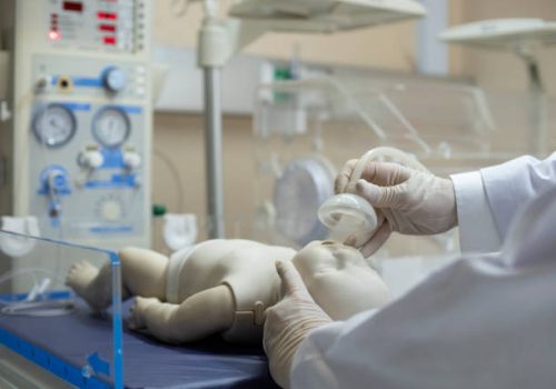 A doctor putting an oxygen mask on a newborn infant model under a radiant warmer/resuscitator unit.